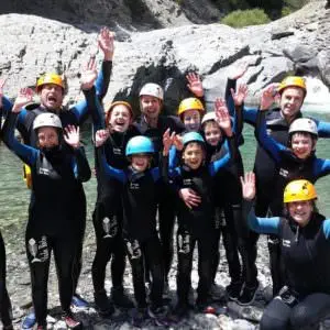 Groupe familial en combinaison néoprène et casque, souriant et levant les bras après une session de canyoning dans une rivière des Pyrénées. Eau turquoise, rochers et nature sauvage en arrière-plan.